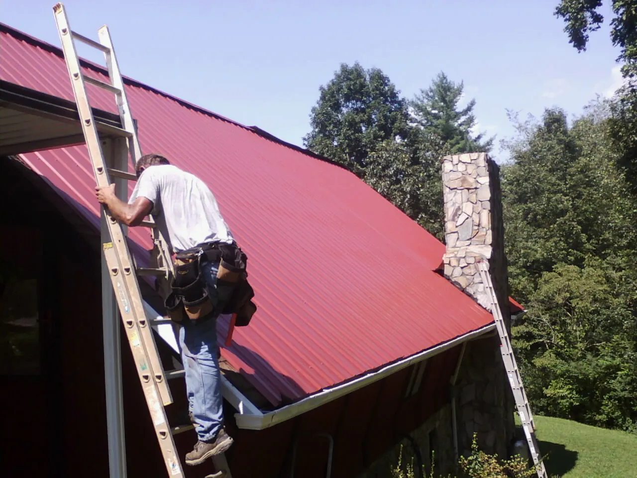 Roofer on a ladder beside a red metal roof and stone chimney — Soffit & Fascia Repair by Roofing Brothers in La Fayette